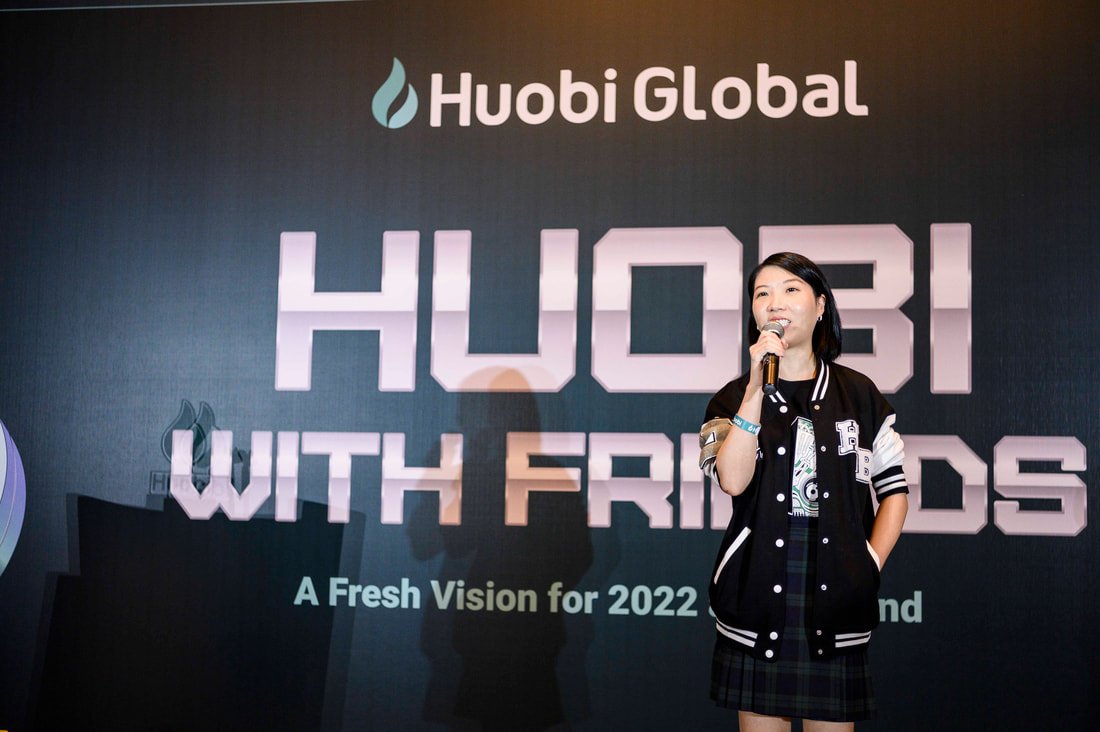 A woman holding a microphone stands in front of a large display screen at a Huobi Global event titled 'Huobi with Friends.' The screen shows the Huobi logo and text that reads 'A Fresh Vision for 2022.'