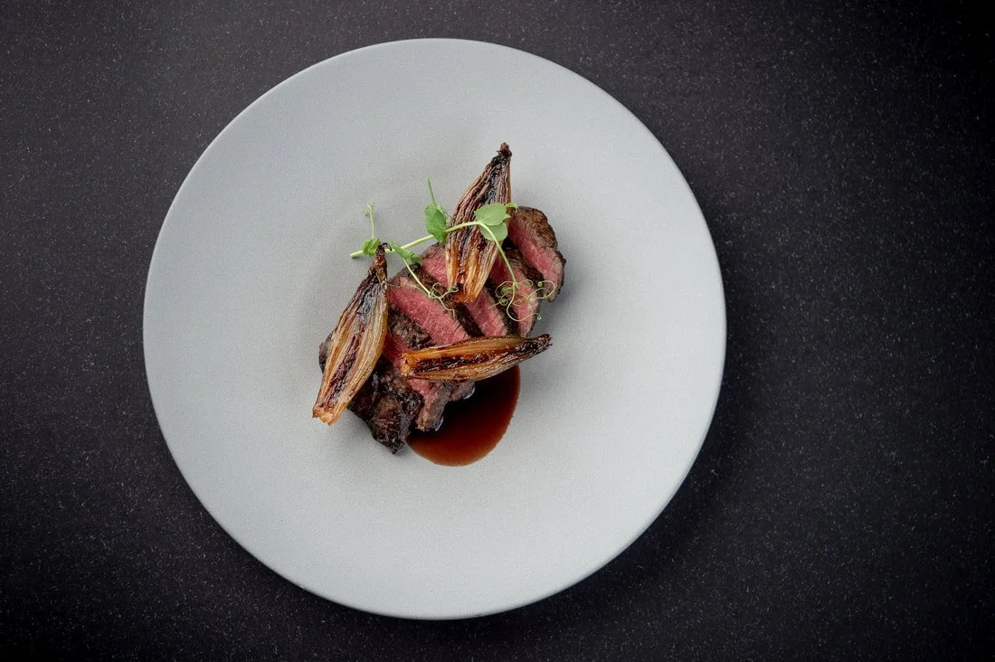 Sliced medium-rare steak with roasted shallots, garnished with microgreens and served with a dark sauce on a white plate.
