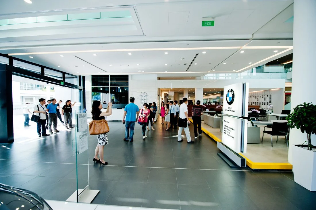 People inside a car dealership, with BMW branding, waiting and walking around, some taking photos, in a modern showroom with large windows and bright lighting.