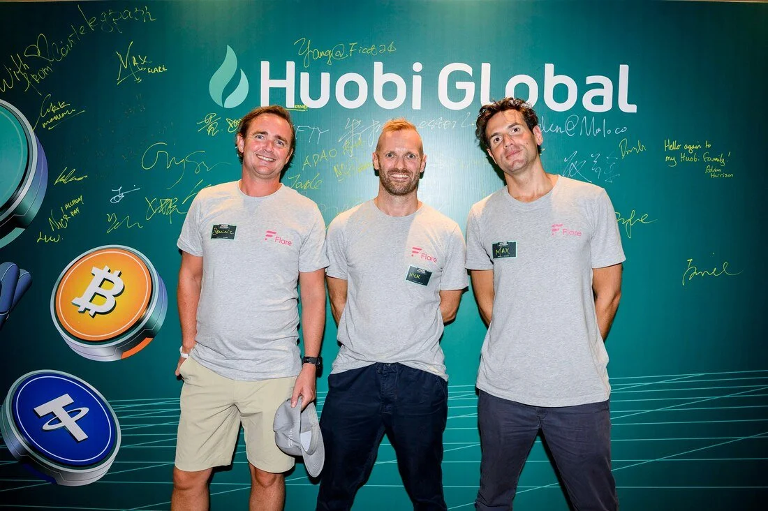 Three men standing in front of a green background with the words 'Huobi Global'. They are wearing gray t-shirts with name tags and are smiling. The background features signatures and graphics of cryptocurrency symbols.