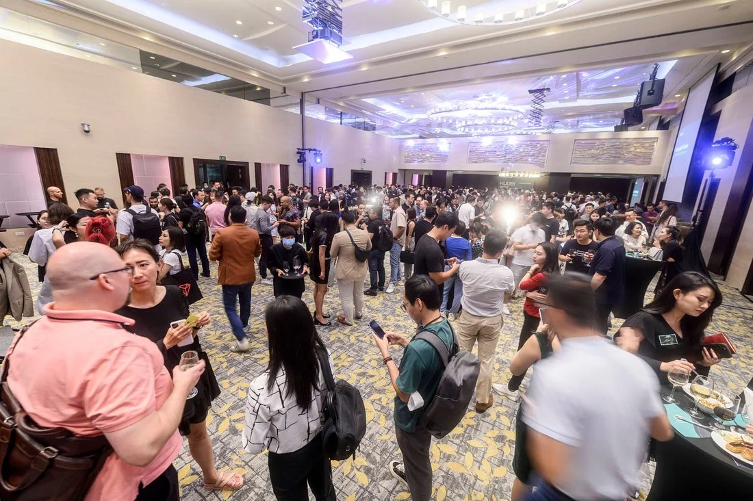 A large group of people attending a conference or event in a spacious, modern ballroom with high ceilings, decorative lighting, and large screens.