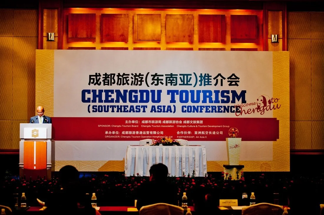A man is speaking at a podium during the Chengdu Tourism (Southeast Asia) Conference, with a large banner behind him displaying event details. The stage has a table with flowers and a small stand with a sign that says "Chengdu". The backdrop indicate