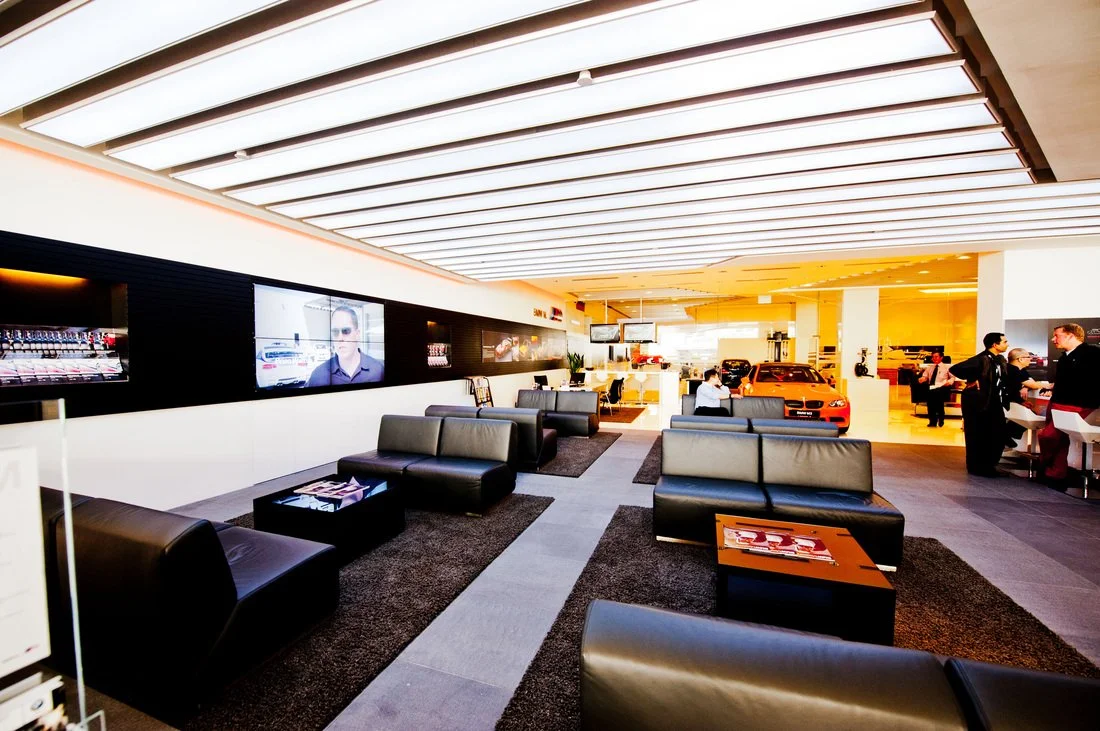 Modern car dealership showroom with black sofas, display TVs, and a bright orange sports car, with customers and staff interacting.