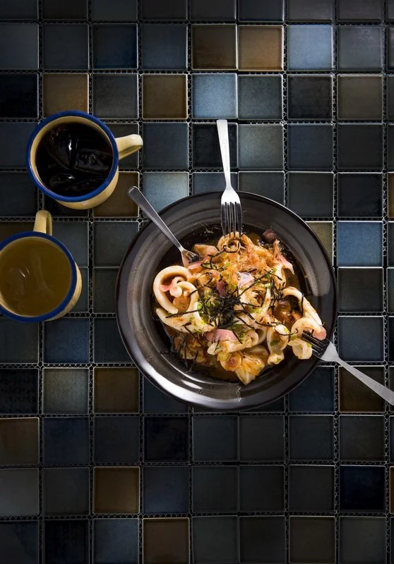 Pasta dish with seafood garnished with herbs, served on a black plate. There are two mugs of coffee, one with a dark liquid and one with a light-colored drink, on a tiled surface.
