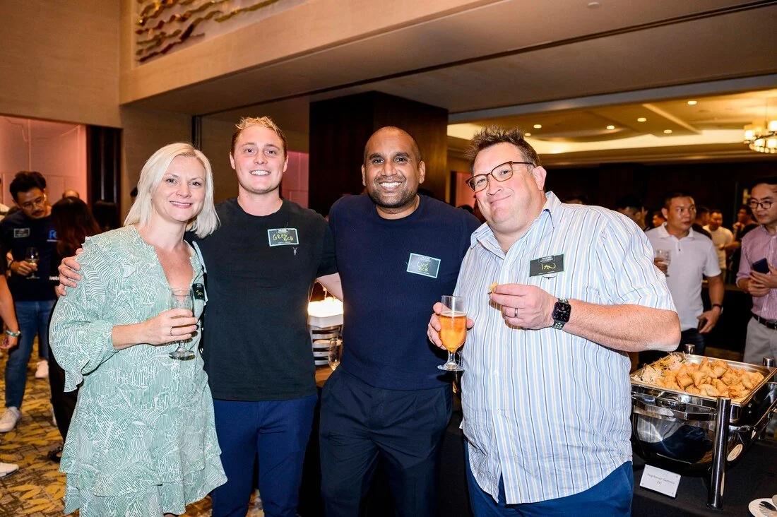 Group of four people smiling and holding drinks at a social event, with others in the background and a buffet table nearby.