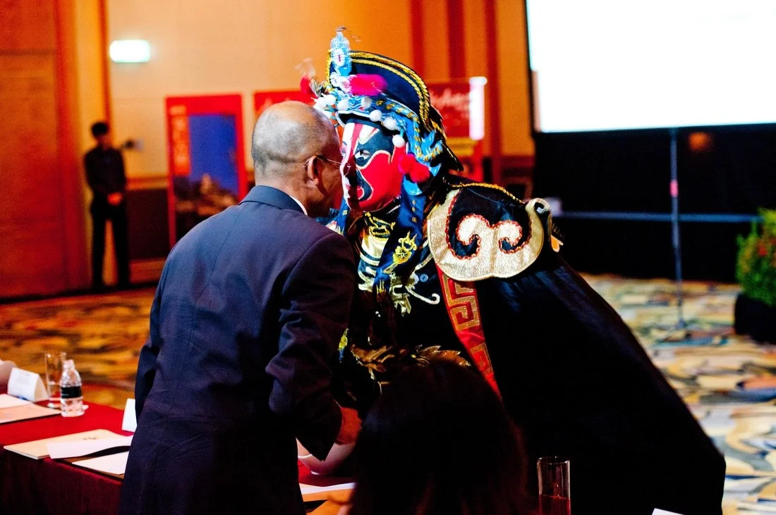 A man in a suit and glasses speaking or greeting a performer in a traditional Chinese opera costume with a painted face at a conference or event.