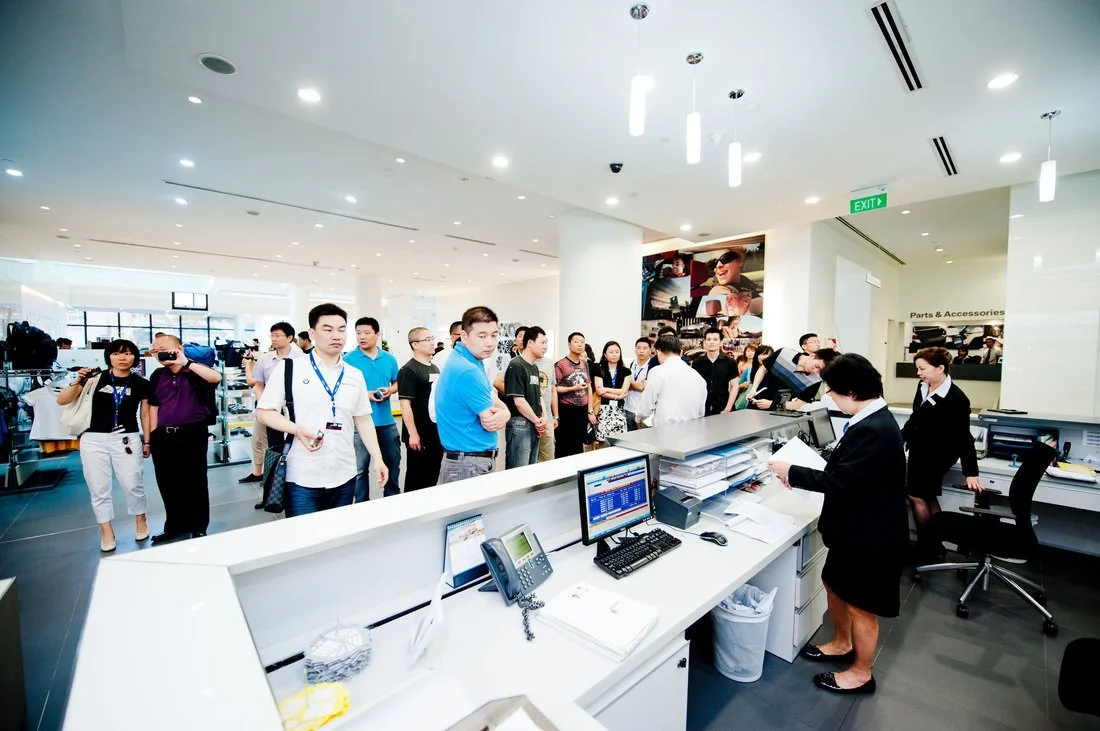 People standing in line at a service counter inside a retail store, with staff behind the counter assisting customers.