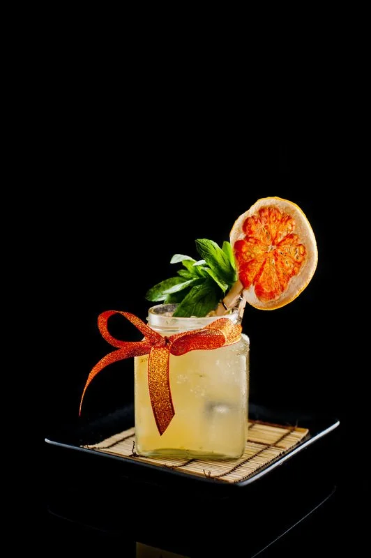 A cocktail in a square glass garnished with a slice of citrus fruit, a sprig of mint, and a ribbon, placed on a black tray against a black background.