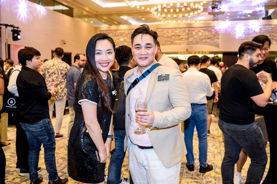 Two people smiling and posing for a photo at a social event, with a crowd of people in the background.
