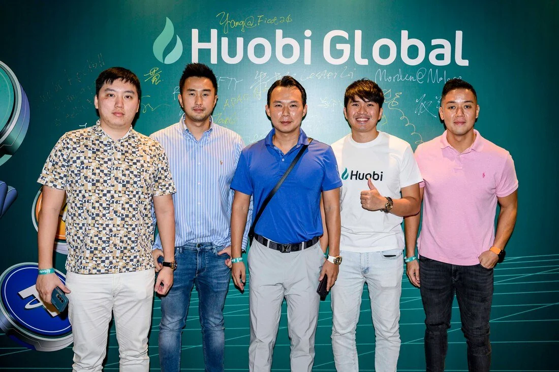 Five men standing in front of a green Huobi Global backdrop, smiling, some giving a thumbs up, dressed in casual and semi-formal attire, with signatures and writings on the backdrop.
