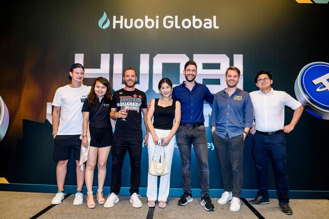 Group of seven people standing together in front of a Huobi Global banner at a conference or event, smiling and posing for a photo.
