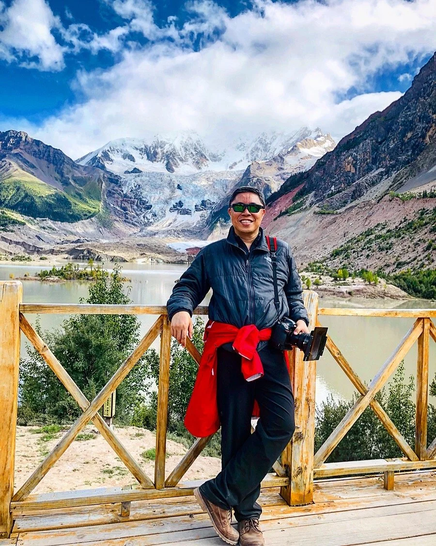 Man standing on a wooden platform with a scenic mountain lake and snow-capped peaks in the background. He is smiling, wearing sunglasses, a black jacket, with a red jacket tied around his waist, and holding a camera.