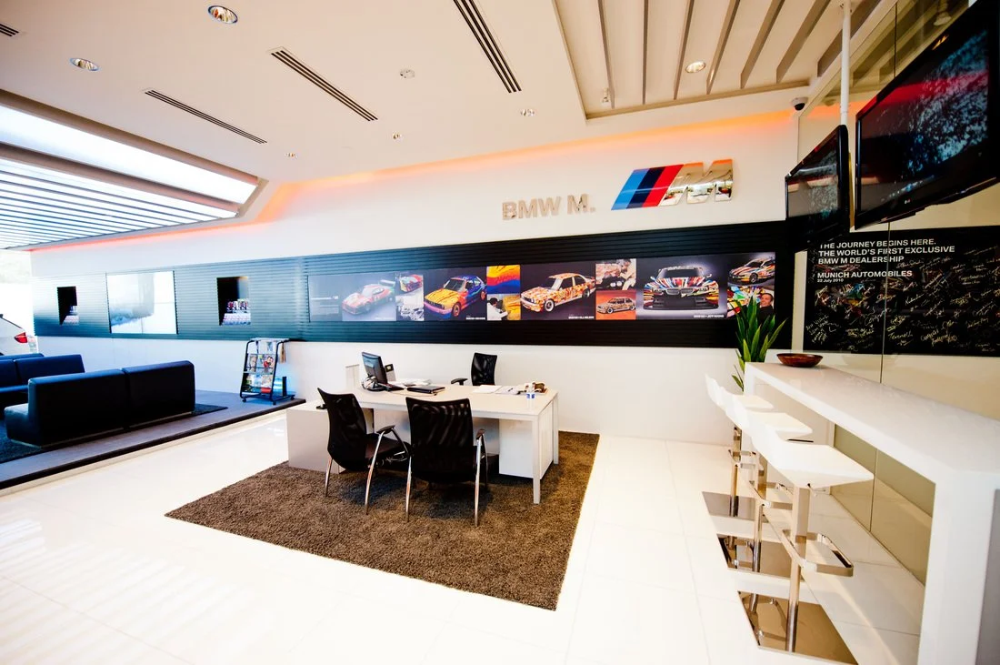 Interior of a BMW dealership meeting room with a white table, four black chairs, and BMW M car images on the wall.