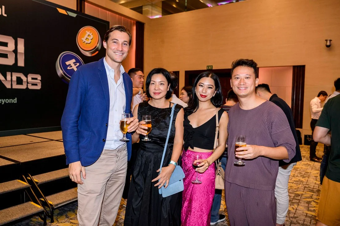 Group of four smiling people holding glasses of champagne at a social event with a Bitcoin-themed backdrop.