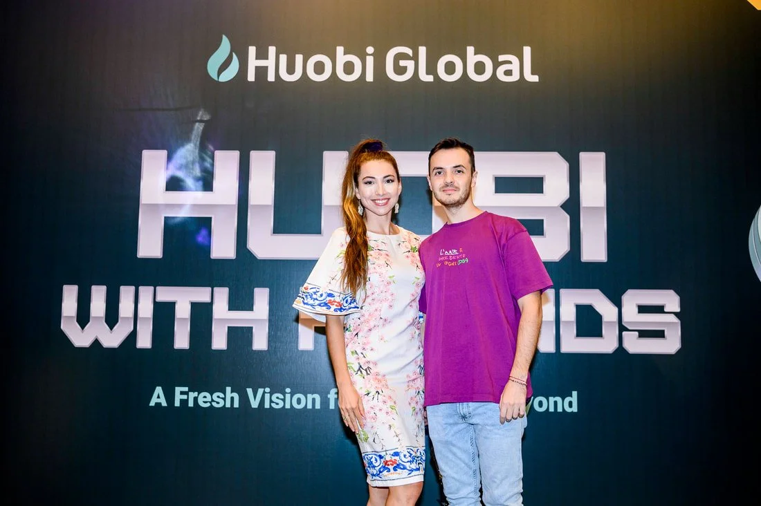 A woman and a man standing together and smiling in front of a dark backdrop with the words 'Huobi Global' and 'HUBI WITH FRIENDS' on it. The woman has long red hair and is wearing a floral dress. The man has short dark hair and is wearing a purple T-