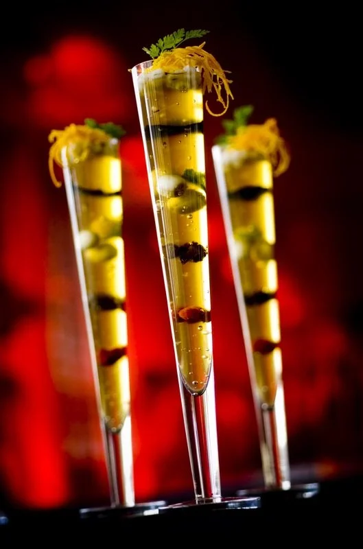 Three tall, skinny glasses filled with yellow liquid, garnished with herbs and shredded yellow garnish, with some berries or fruit pieces inside, set against a blurred red and black background.