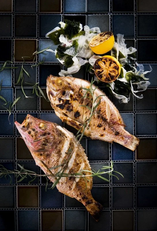 Two grilled fish on a black tiled surface, garnished with rosemary, along with lemon halves, some greens, and white flowers.