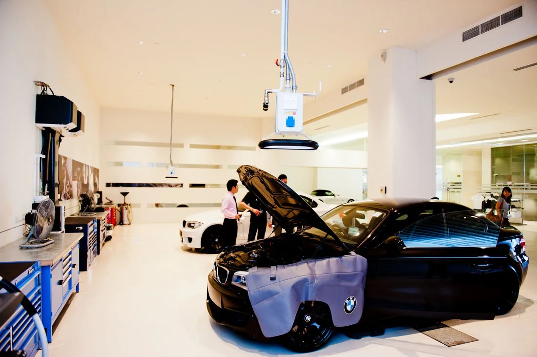 A sleek black convertible car with a BMW logo has a partial hood cover and is parked inside a spacious, modern car service center. Other cars and a mechanic are visible in the background within the well-lit, white interior of the facility.