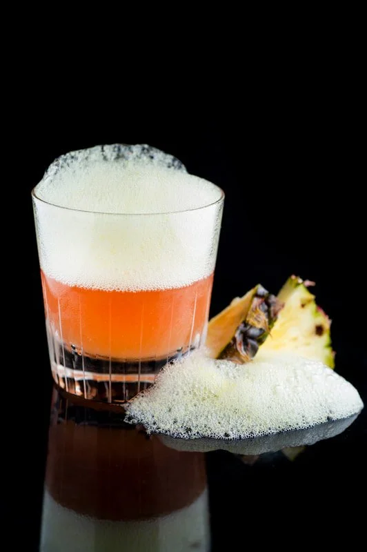 A cocktail with foam in a glass, pineapple slices, and foamy bubbles on a black reflective surface.