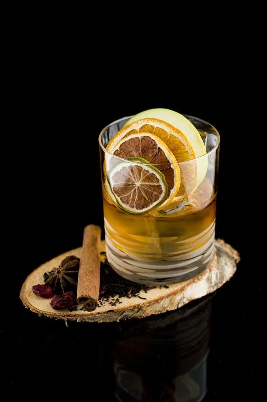 A glass of iced tea with lemon, orange, and lime slices, garnished with cinnamon sticks, star anise, and dried berries on a wooden slice with black background.