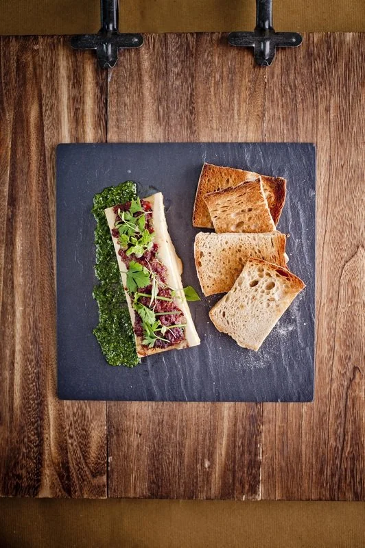 A slate board with a piece of toast topped with cheese, herbs, and greens, alongside three pieces of toasted bread, on a wooden table.