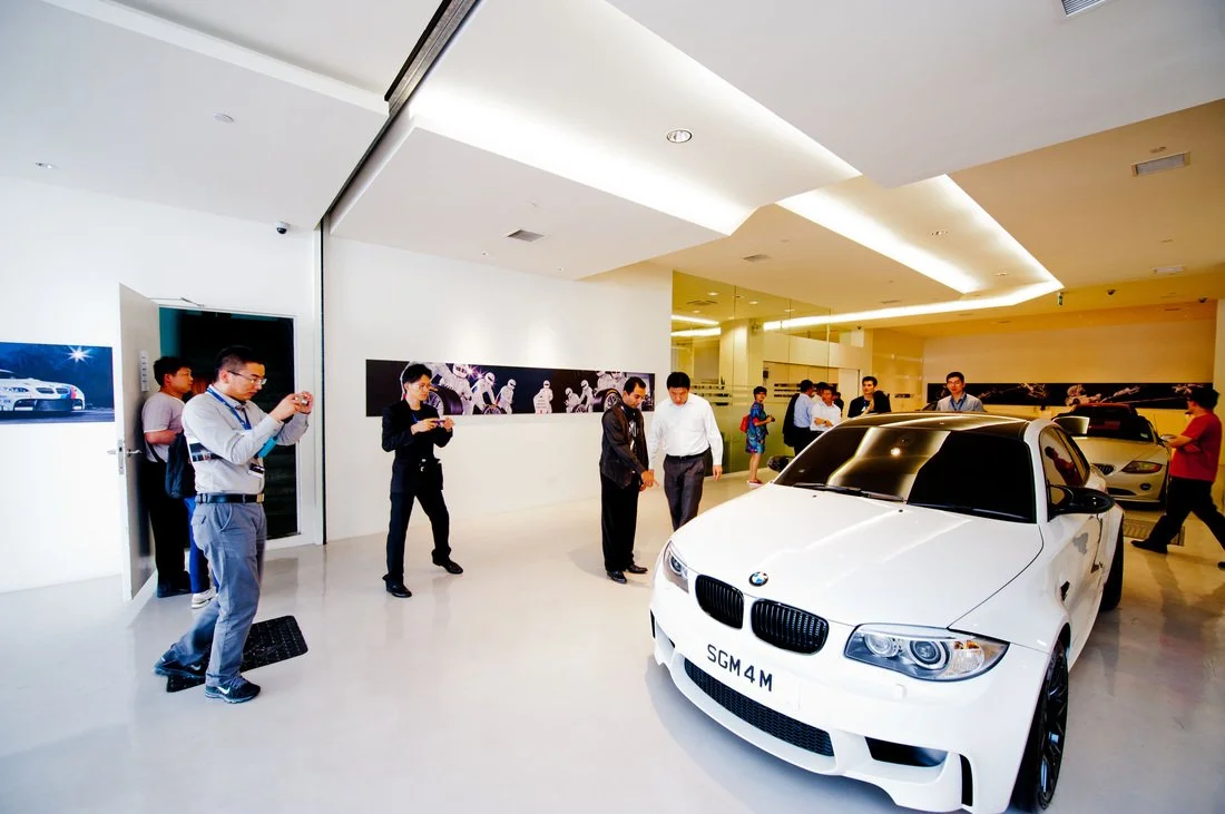 White BMW car displayed in a showroom with people around, some taking photos.