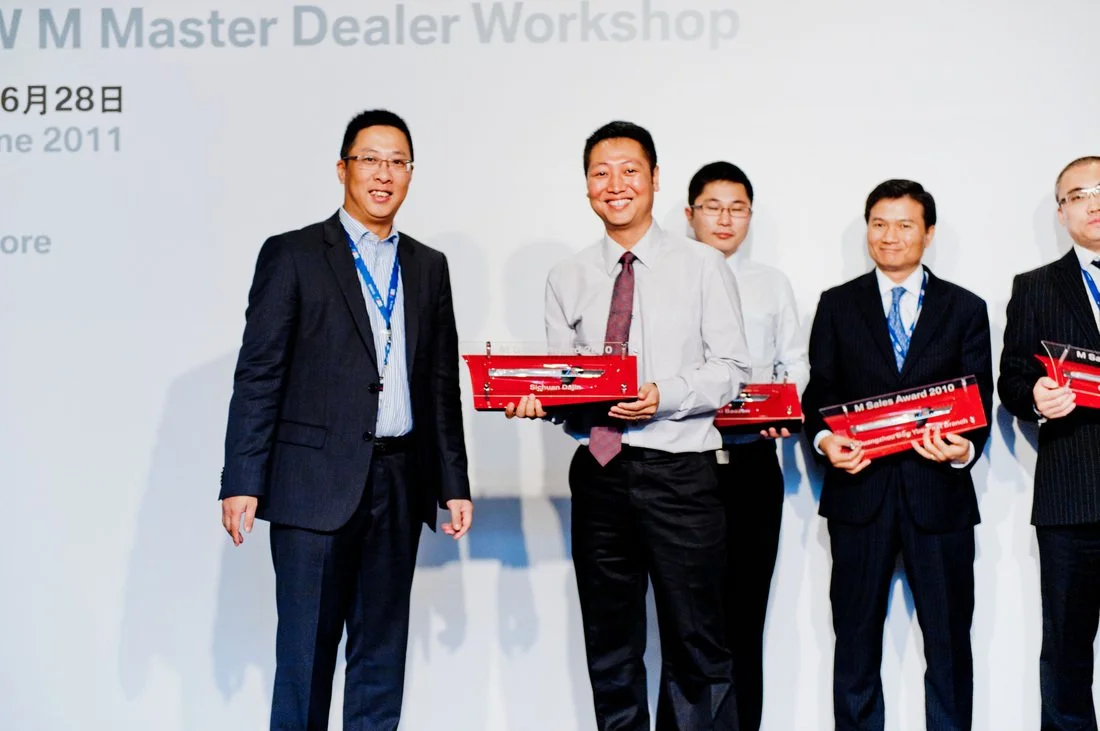 Group of men in formal suits standing on stage during an award ceremony, some holding red awards.