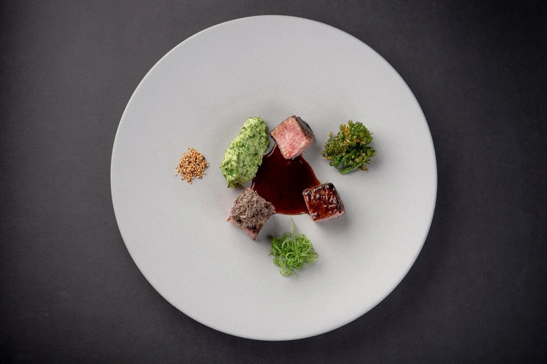 Elegant white plate with small portions of gourmet food, including pieces of meat, a green mashed item, a green garnish, and a red sauce.
