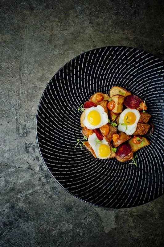 A black textured bowl containing roasted potatoes, carrots, and small tomatoes, topped with three soft-boiled eggs garnished with green herbs, placed on a dark gray concrete surface.