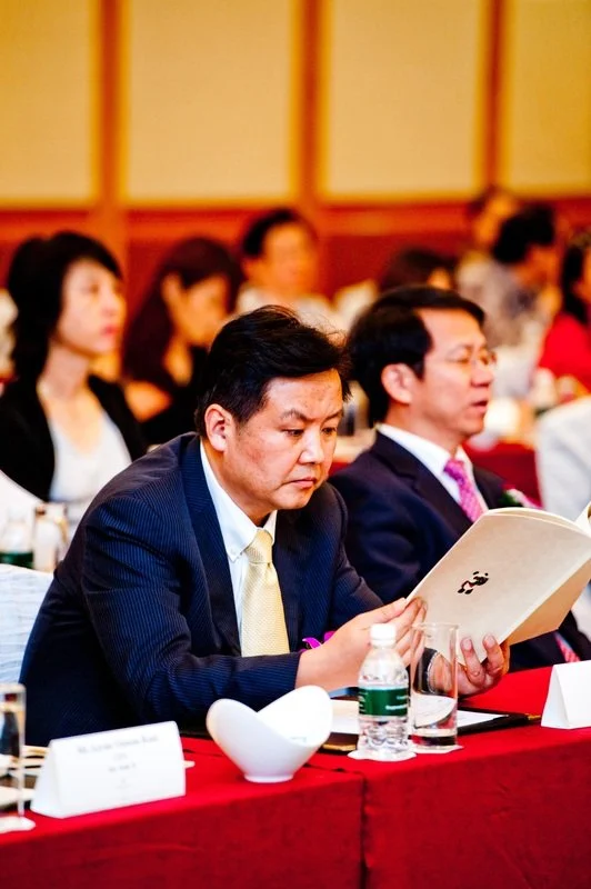 A man in a dark suit and yellow tie reading a booklet at a conference table with others seated behind him.