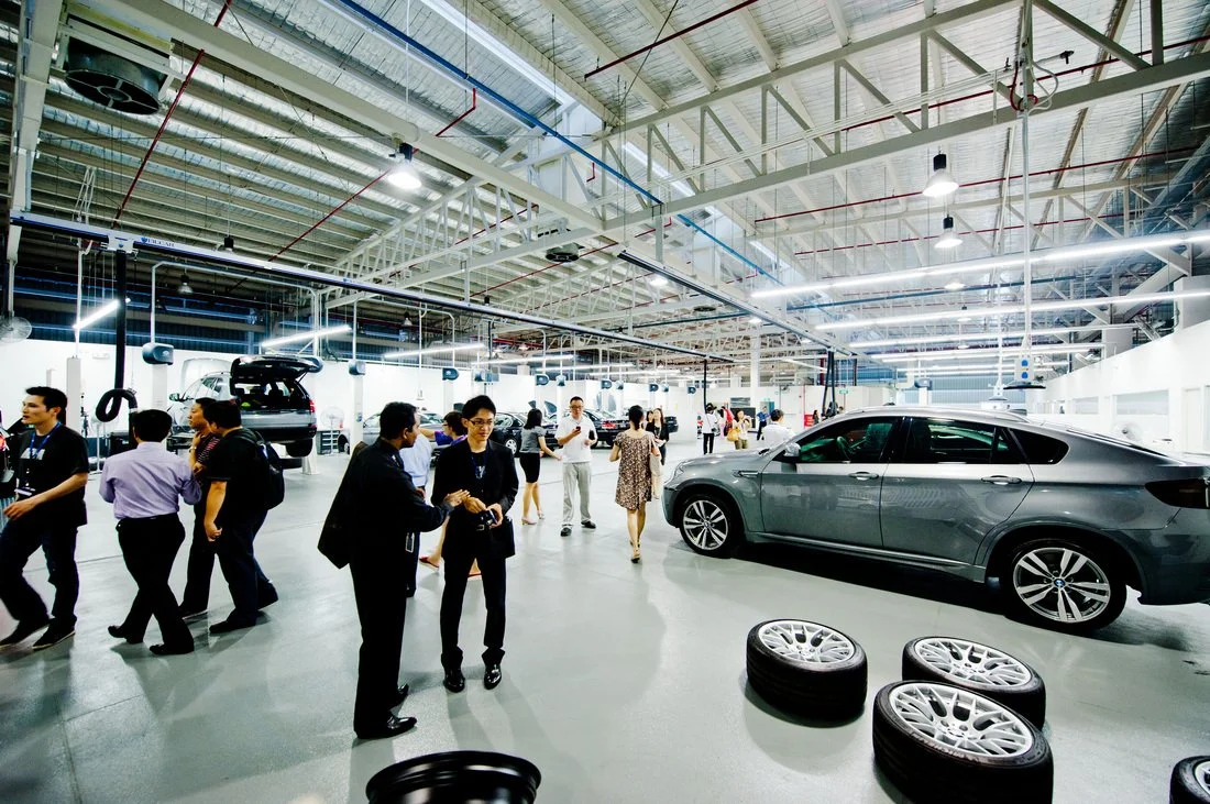 An indoor automotive manufacturing or assembly plant with cars on display, workers and visitors walking around, and tires on the floor.