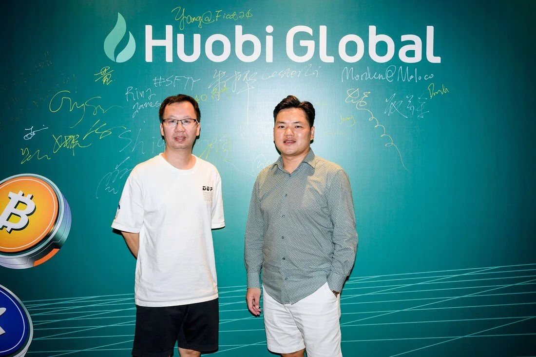 Two men standing in front of a teal background with the text "Huobi Global" and the Huobi logo, along with signatures and writings. One man is wearing glasses and a white T-shirt, the other is in a gray shirt and white shorts.
