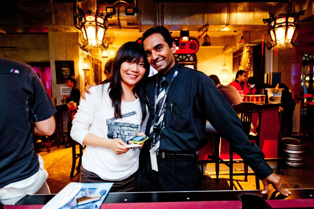A woman and a man smiling and posing together in a restaurant or bar, with warm lighting and decorative lanterns hanging from the ceiling in the background.