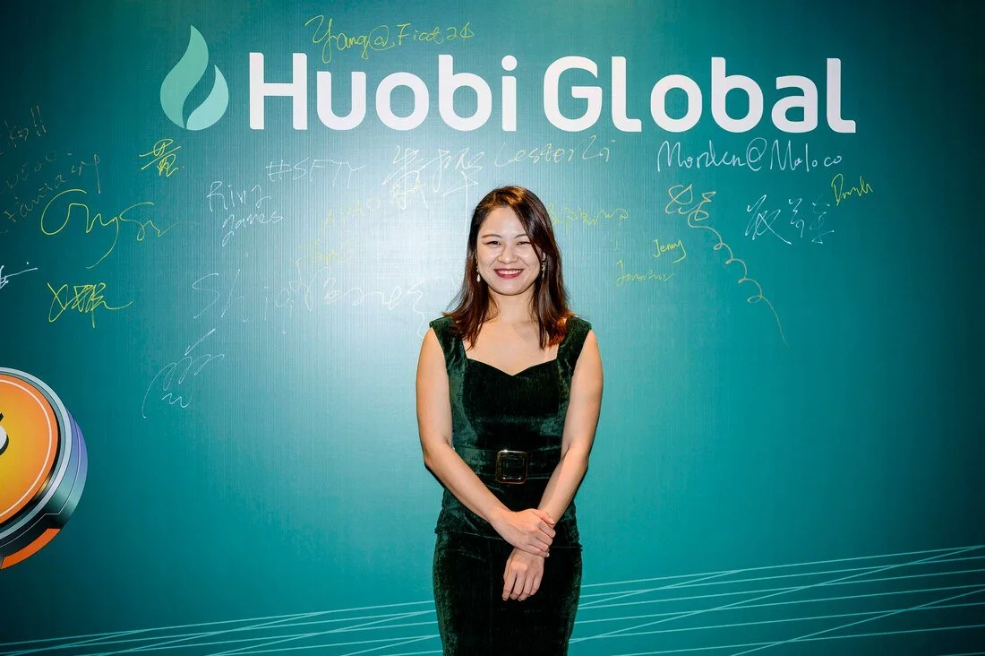 A woman in a dark green velvety dress standing in front of a teal wall with 'Huobi Global' logo, signatures, and handwritten notes.