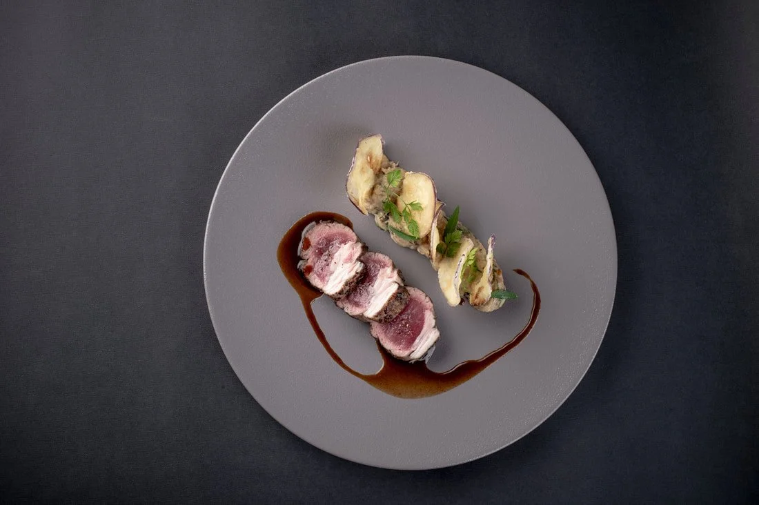 A plate with sliced, cooked meat topped with sauce, garnished with herbs, and a side of grilled vegetables on a dark background.
