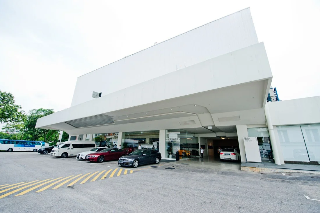 Modern white building with a parking area in front, cars parked outside, and a few people near the entrance.