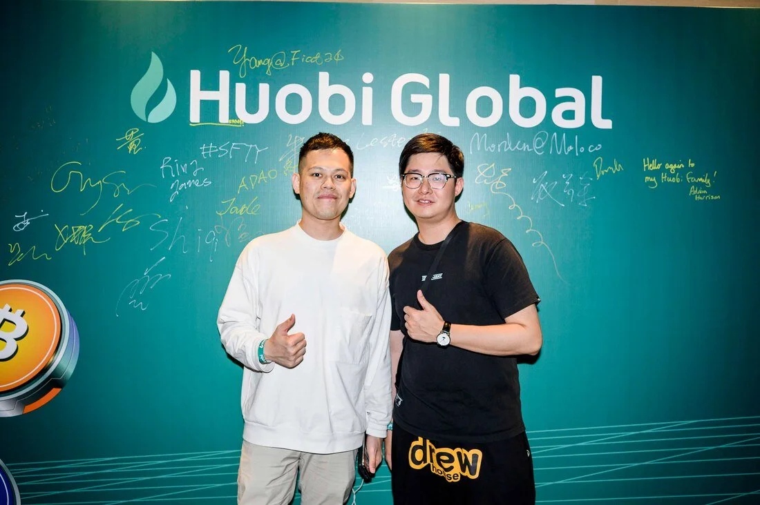 Two men standing in front of a teal-colored background with the words 'Huobi Global' and a flame logo. Both are giving a thumbs-up. The man on the left is wearing a white long-sleeve shirt and beige pants, while the man on the right is wearing a blac