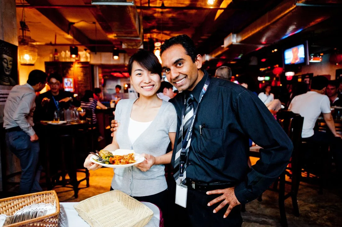 A smiling woman holding a plate of food with a man standing next to her in a lively restaurant or bar.