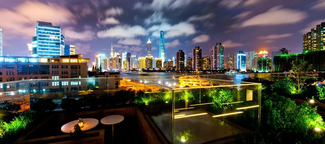 Nighttime city skyline with illuminated skyscrapers and clouds in the sky, viewed from a rooftop with outdoor seating and greenery.