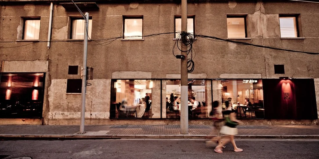 People sitting and dining inside a restaurant viewed through large glass windows at night, with an old building exterior and two blurred pedestrians walking by on the sidewalk.