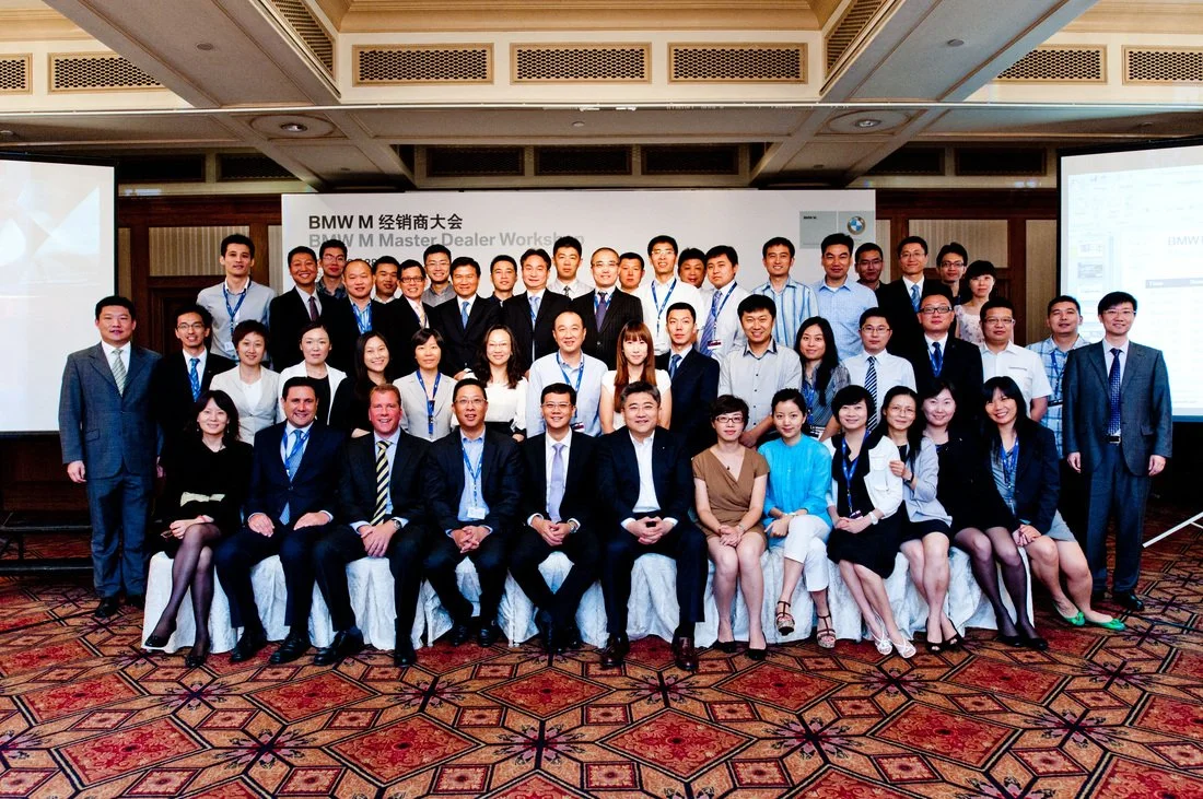 Group of professionals at a BMW event, posing for a photo in a conference room with a banner and presentation screens.