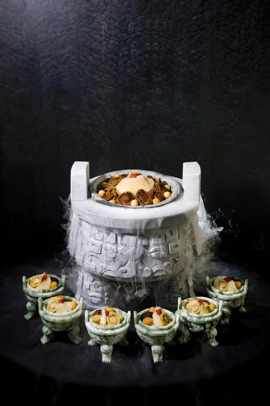 Large white stone altar with steam rising from it, surrounded by six smaller stone bowls filled with food and slices of fruit, set against a black background.