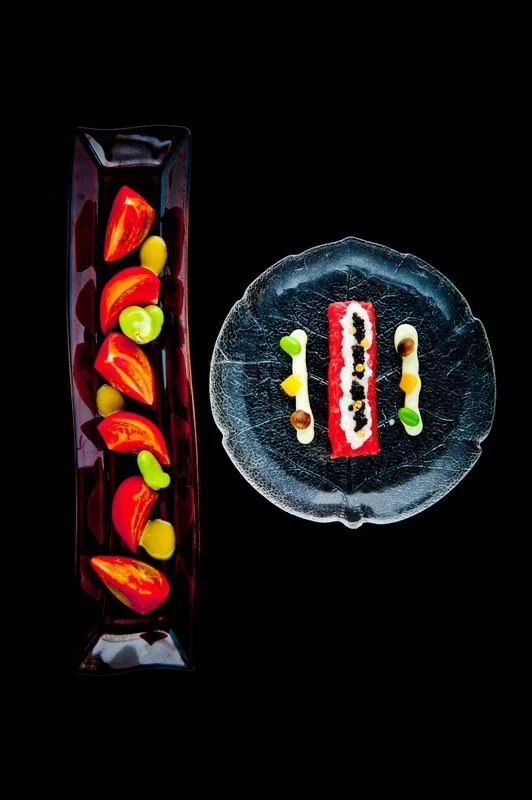 Artistic desserts on black background, including a tall rectangular glass with red and yellow fruit slices, and a round textured glass plate with a red and white layered dessert topped with black seeds, accompanied by small green, yellow, and brown dollops.