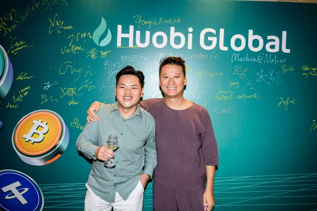 Two men smiling, one holding a wine glass, standing in front of a blue-green wall with 'Huobi Global' logo and handwritten signatures or notes, with large Bitcoin and other cryptocurrency symbols.