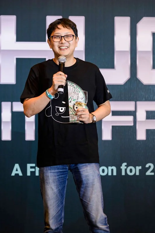 Smiling man with glasses holding a microphone and a trophy on stage at an event.