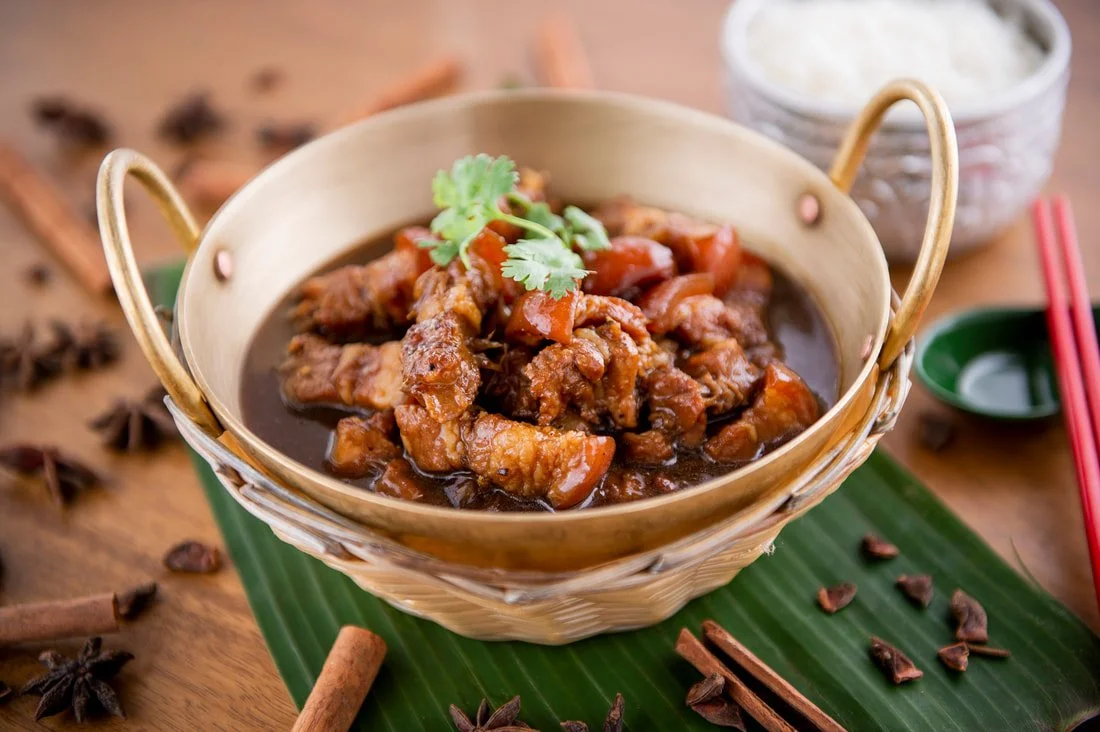 Bowl of beef stew garnished with cilantro, served in a traditional woven basket on a green banana leaf, with cinnamon sticks, star anise, and chopsticks nearby.