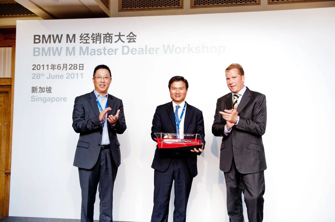 Three men in suits standing on a stage with a white backdrop that has text in English and Chinese, during a BMW M Master Dealer Workshop event on June 28, 2011, in Singapore. The men are holding a red model car.