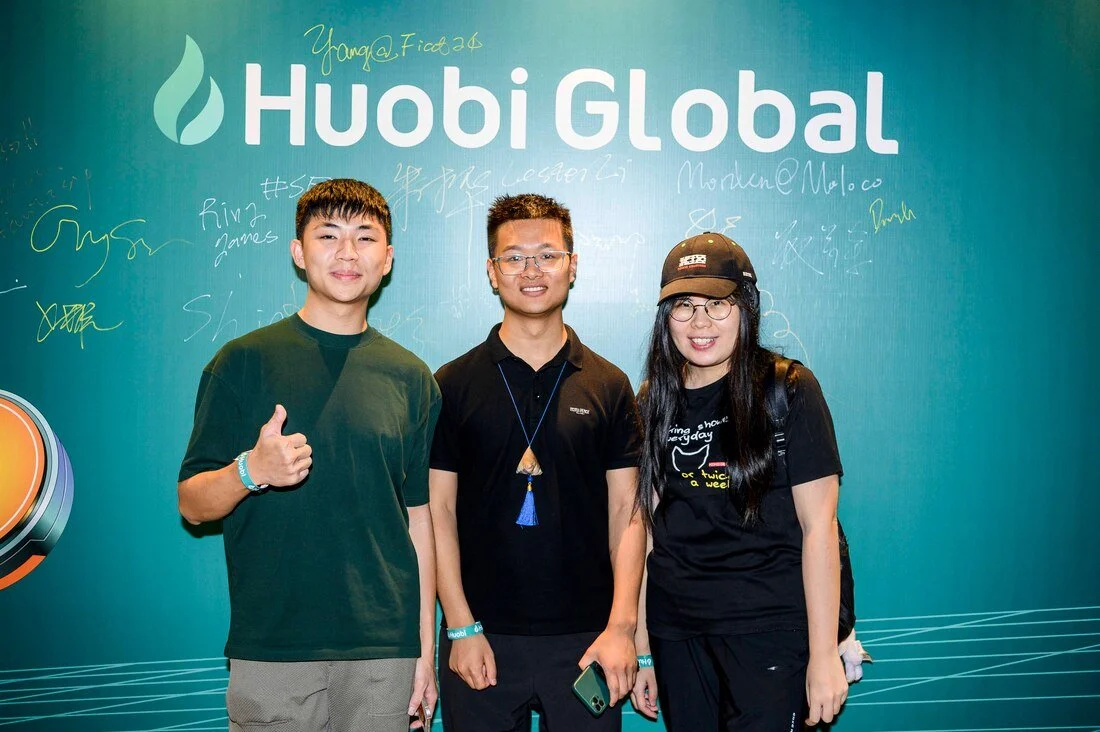 Three people standing in front of a Huobi Global sign with signatures, smiling at the camera.