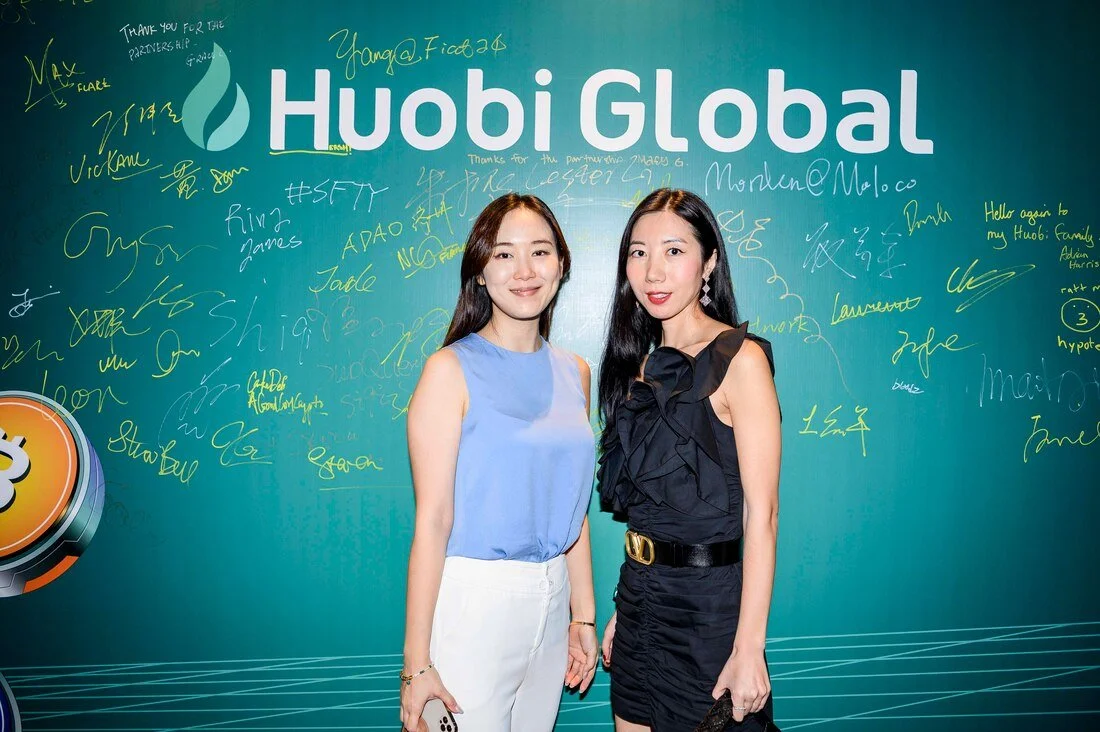 Two women standing in front of a green Huobi Global backdrop with signatures and messages in various colors. One woman wears a light blue sleeveless top and white pants, the other a black dress with ruffled details and a gold belt.