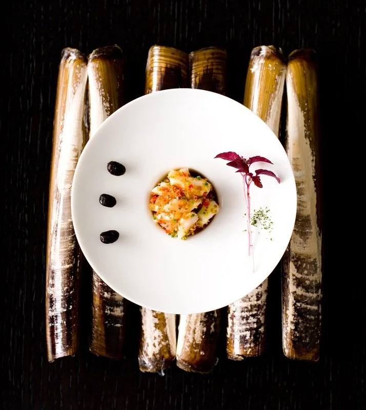 A white plate with a small portion of colorful seafood or vegetable dish, three black dots, and a purple herb garnish, placed on a dark wooden surface with five bamboo stalks in the background.